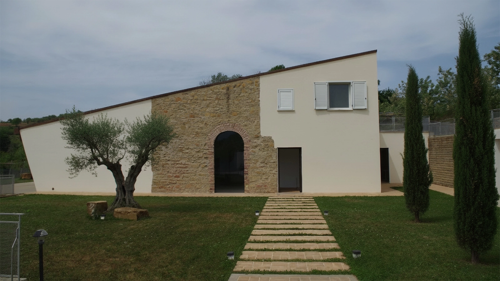 Facciata esterna del Casale Coni con ingresso e portale originale ad arco in pietra e mattoni, superfici intonacate e giardino con percorso pedonale. Vegetazione mediterranea composta di olivi e cipressi.