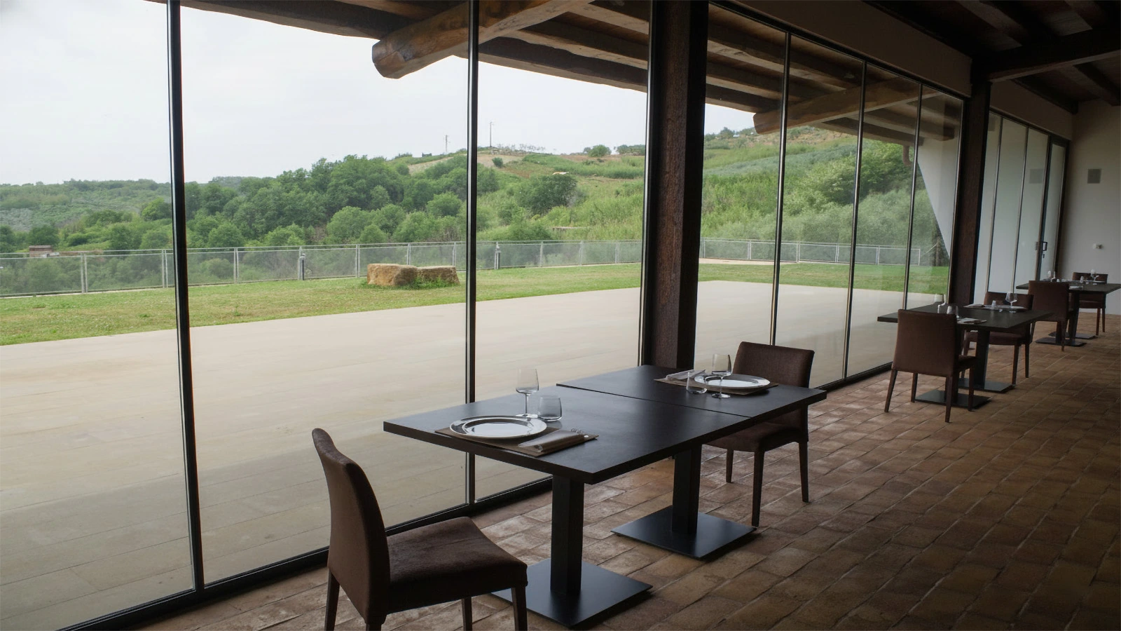 Sala di ristorazione del Casale Coni con ampia vetrata panoramica affacciata sul paesaggio rurale; interno caratterizzato da soffitto con travi in legno a vista, pavimentazione in cotto e tavoli apparecchiati.